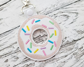 May include: White donut-shaped keychain with pink frosting and colorful sprinkles. The keychain has a silver metal ring.