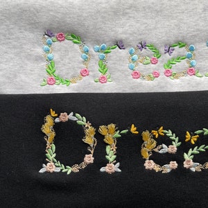 May include: The word "Dream" is embroidered in colourful flowers and leaves on a white and black sweatshirt. The flowers are pink, blue, and yellow.