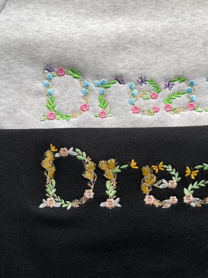 May include: The word "Dream" is embroidered in colourful flowers and leaves on a white and black sweatshirt. The flowers are pink, blue, and yellow.
