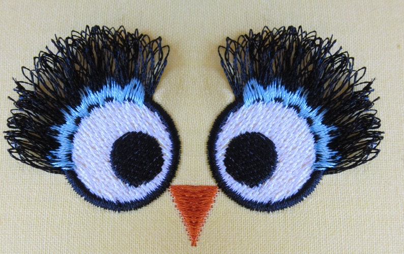 May include: Embroidered owl eyes with black eyelashes, blue eyelids, white pupils, and black centers. The owl has a small orange beak.