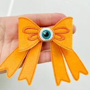 May include: An orange felt bow with an embroidered eye in the center. The bow has two long, pointed tails and orange stitching around the edges. The eye is white, teal, and black.