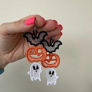 May include: A pair of Halloween-themed earrings featuring black bats, orange pumpkins, and white ghosts. The earrings are made of delicate lace and are perfect for adding a touch of spooky fun to any outfit.