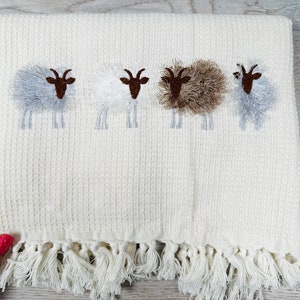 May include: White waffle weave towel with a fringe border and four embroidered sheep with brown faces and fluffy white, gray, and brown wool.