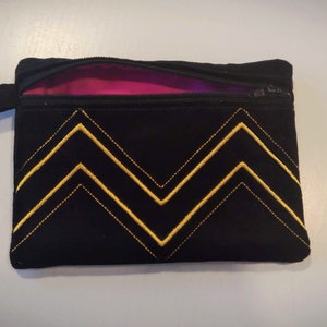 Può includere: Una pochette nera con cerniera, fodera rosa e un motivo a chevron dorati.