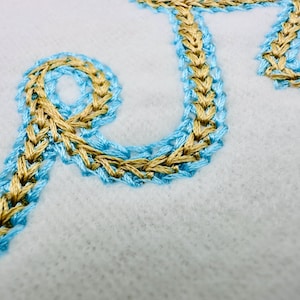 Puede incluir: Primer plano de texto bordado en hilo dorado y azul claro sobre tela blanca. La costura tiene una apariencia trenzada. El dise&ntilde;o es probablemente parte de un proyecto textil m&aacute;s grande.