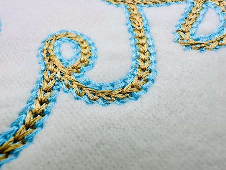 Puede incluir: Primer plano de texto bordado en hilo dorado y azul claro sobre tela blanca. La costura tiene una apariencia trenzada. El dise&ntilde;o es probablemente parte de un proyecto textil m&aacute;s grande.
