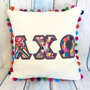 Puede incluir: Una almohada blanca con un ribete de pompones de colores. La almohada tiene las letras "AXO" cosidas en ella en un estilo patchwork.
