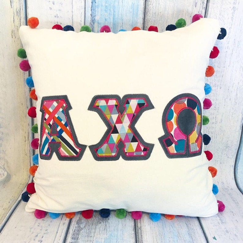 Puede incluir: Una almohada blanca con un ribete de pompones de colores. La almohada tiene las letras "AXO" cosidas en ella en un estilo patchwork.