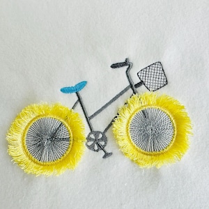 May include: Embroidered bicycle design with yellow fringed wheels, a blue seat, and a black basket. The bicycle frame is gray, set against a white background. This design is suitable for various craft projects.