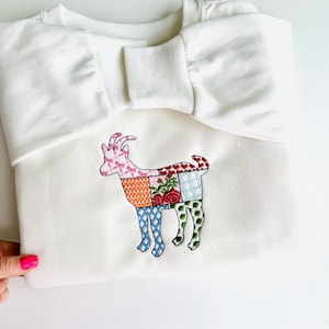 Puede incluir: Sudadera blanca con un diseño de cabra de patchwork bordado en la parte delantera. La cabra está hecha de cuadrados de diferentes colores con patrones florales y geométricos.