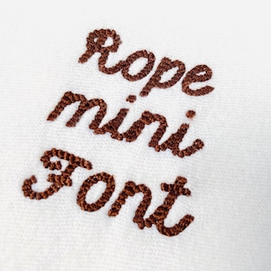 Puede incluir: Primer plano de tela blanca con texto bordado en marrón. El texto dice "Rope mini Font" en un estilo cursivo. El bordado tiene una apariencia texturizada, similar a una cuerda.