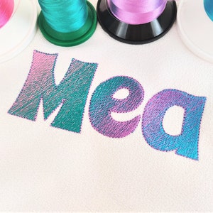 Może przedstawiać: Wyhaftowane imię "Mea" nicią różową i turkusową na białym tle tkaniny. Imię jest wyszyte pogrubioną, prostokątną czcionką.