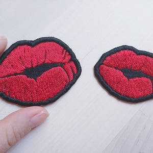 以下が含まれることがあります： 黒い輪郭のある赤い刺繍の唇のパッチが2つ。大きなパッチは左側にあり、小さなパッチは右側にあります。