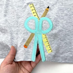 May include: A light gray fabric featuring an embroidered applique design of a pair of light blue scissors layered over a yellow ruler and a yellow pencil with a black lead tip. The ruler displays black markings and numbers from 1 to 4.