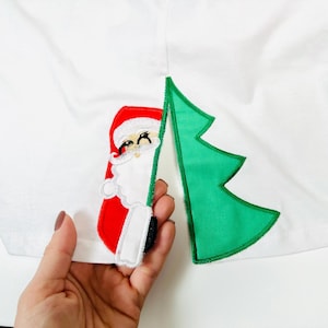 Pode incluir: Uma camiseta branca com uma aplicação de Papai Noel vermelha e branca e uma aplicação de árvore de Natal verde.