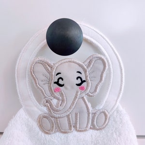 May include: White towel with a grey embroidered elephant with pink cheeks. The towel has a white ring sewn around the edge and is hanging on a black hook.