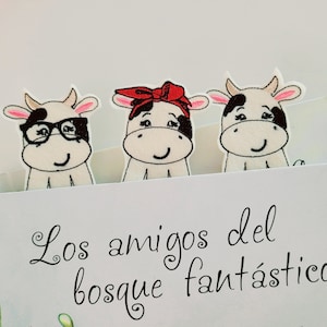 May include: Three embroidered cow bookmarks with black and white spots. The cow in the middle has a red headband. The text on the book reads "Los amigos del bosque fantástico".