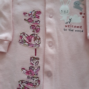 May include: A light pink baby onesie with the name "Baby" embroidered in a floral pattern. The onesie features a graphic of a white bunny, a gray bunny, and the text "welcome to the world".