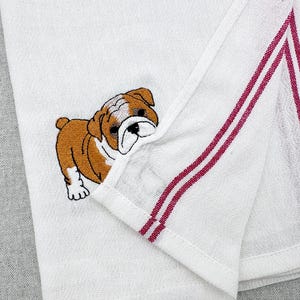 Pode incluir: Toalha de cozinha branca com um desenho de bulldog castanho e branco bordado. A toalha tem duas riscas bordô ao longo da borda. A toalha está dobrada, mostrando o bulldog e as riscas.