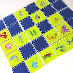 Könnte beinhalten: Ein Set mit 24 Filzquadraten, auf denen verschiedene Meeresbewohner und Objekte gestickt sind. Die Quadrate sind gelb mit blauen Nähten und die Bilder sind in verschiedenen Farben gehalten. Die Bilder zeigen ein Segelboot, einen Seestern, einen Oktopus, eine Krabbe, eine Muschel, einen Delfin, einen Fisch, eine Schildkröte und einen Anker.