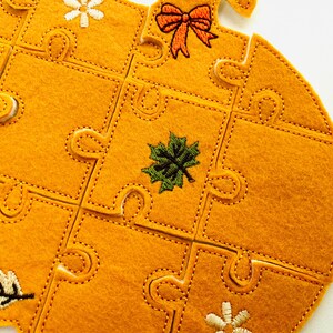 May include: A felt puzzle in the shape of a pumpkin, with a bright orange color. The puzzle pieces are stitched together with a contrasting thread. The pumpkin is decorated with embroidered fall-themed elements, including a bow, leaves, and flowers.