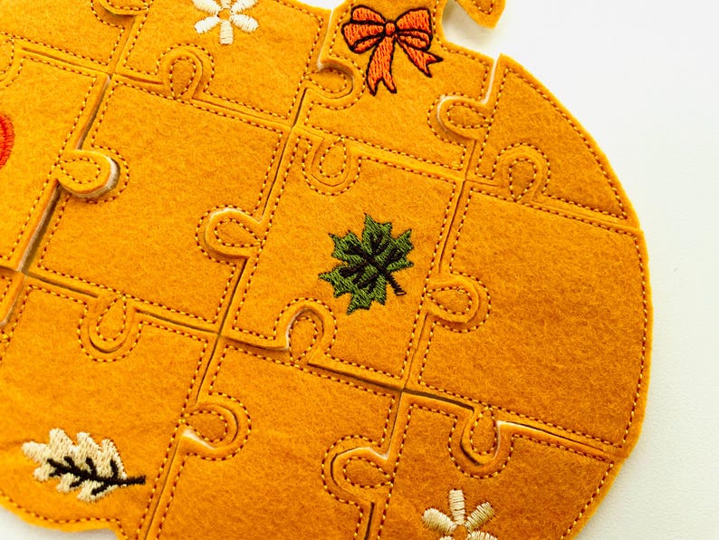 May include: A felt puzzle in the shape of a pumpkin, with a bright orange color. The puzzle pieces are stitched together with a contrasting thread. The pumpkin is decorated with embroidered fall-themed elements, including a bow, leaves, and flowers.