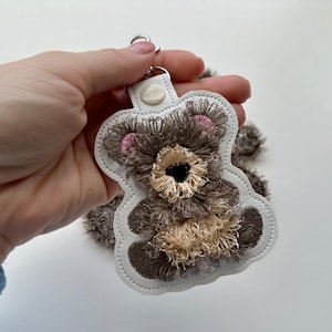 Op de afbeelding: Een witte vilt sleutelhanger met een bruine en beige geborduurde teddybeer. De beer heeft roze oren en een zwarte neus.