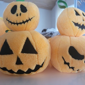 May include: Three plush pumpkin toys with different facial expressions. The pumpkins are orange and have black eyes, noses, and mouths. The pumpkins are stacked on top of each other.