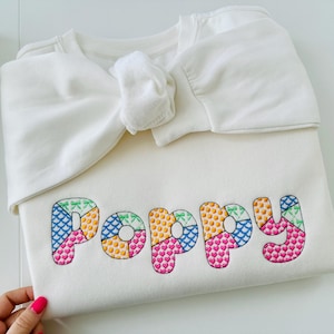 May include: White sweatshirt with a bow on the top and the name "Poppy" embroidered in colorful patchwork letters.