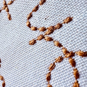 May include: Close-up of a white fabric with a brown embroidered design. The design is made up of a series of interlocking circles and loops.
