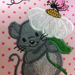 May include: A gray embroidered mouse holding a white flower with a yellow center and a pink butterfly on top. The background is pink with white polka dots.