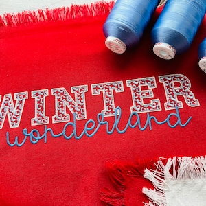 May include: A red fabric with the embroidered text "WINTER wonderland" in white and blue. Three spools of blue thread are in the background. The fabric has a fringed edge, suggesting a decorative item.