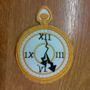 May include: A gold-colored pocket watch with a white face and black Roman numeral markings. The hands of the watch are pointing to the number 11 and the number 7.