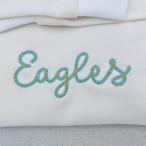 Puede incluir: Tela color crema con la palabra "Eagles" bordada en cursiva. El bordado est&aacute; delineado en azul claro con un relleno dorado. Una cinta blanca es visible en la parte superior.