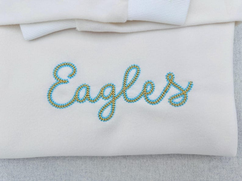 Puede incluir: Tela color crema con la palabra "Eagles" bordada en cursiva. El bordado est&aacute; delineado en azul claro con un relleno dorado. Una cinta blanca es visible en la parte superior.