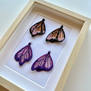 May include: Four embroidered butterfly earrings in a white frame. Two earrings are orange and pink, and two are purple and pink.