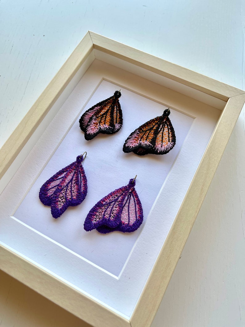 May include: Four embroidered butterfly earrings in a white frame. Two earrings are orange and pink, and two are purple and pink.