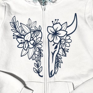 Puede incluir: Sudadera blanca con cremallera y un diseño floral y calavera negro bordado en la parte delantera.