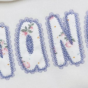 Puede incluir: Primer plano de la palabra "NONO" bordada sobre tela blanca. Las letras están rellenas con un estampado floral en rosa y verde, delineadas con un ribete festoneado azul. El fondo es blanco liso.