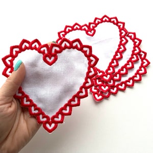 May include: Set of five white heart-shaped fabric coasters with red embroidered edges. Each coaster features a scalloped edge with a repeating heart pattern.