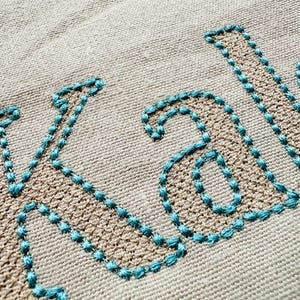 May include: Embroidered name "Kade" in a teal blue thread on a beige linen fabric.