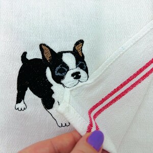 May include: White hand towel with a black and white embroidered Boston Terrier dog. The towel has red stripes along the edge.