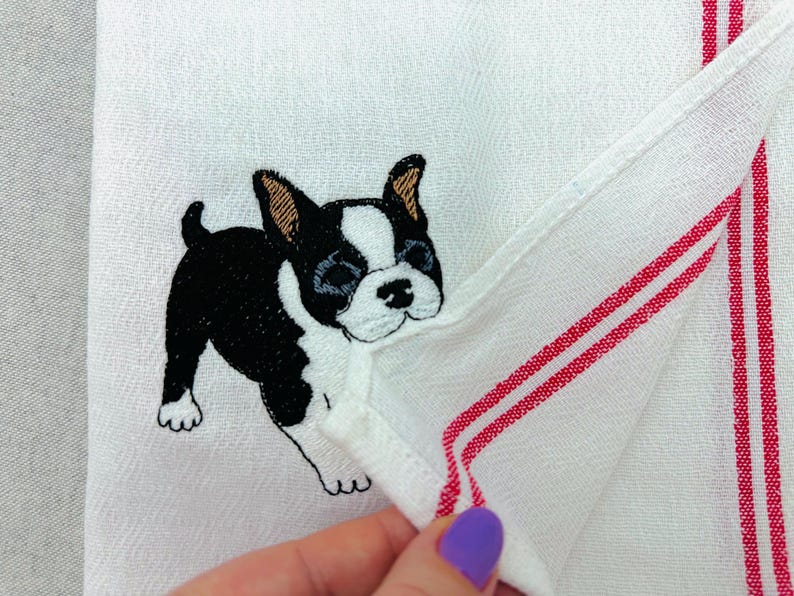 May include: White hand towel with a black and white embroidered Boston Terrier dog. The towel has red stripes along the edge.