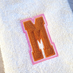 May include: White towel with a large orange letter M embroidered on it. The letter has a pink outline.