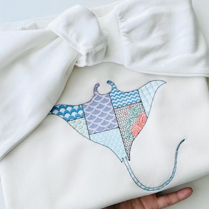 May include: A white sweatshirt with a blue and purple embroidered manta ray design. The manta ray is made up of patchwork squares with different patterns, including a floral design.