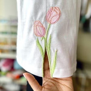 以下が含まれることがあります： 白い生地に、緑の茎と葉を持つ2つのピンクのチューリップの刺繍デザインが施されています。 花の刺繍は詳細で、生地の前面に表示されます。 生地は手で持たれています。