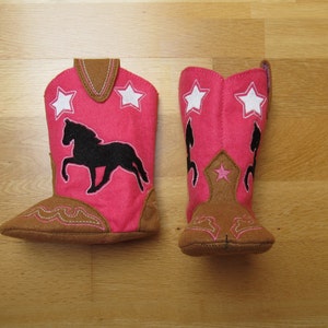 May include: A pair of pink felt cowboy boots with brown felt trim. One boot features a black silhouette of a horse and white stars. The other boot features a brown star.