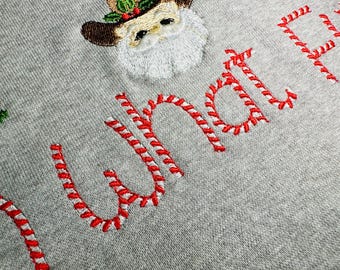 Könnte beinhalten: Ein grauer Pullover mit roter und weißer Zuckerstangen-Stickerei, auf dem "What the Elf" steht.