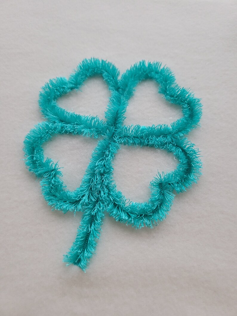 May include: A turquoise four-leaf clover embroidered on a white fabric background.