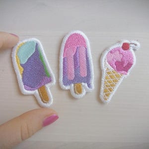 Op de afbeelding: Drie geborduurde strijkpatches met ijsjes. Eén patch is een paarse, roze en groene ijslolly, een andere is een roze en paarse ijslolly en de laatste is een roze ijshoorn met een kers erop.